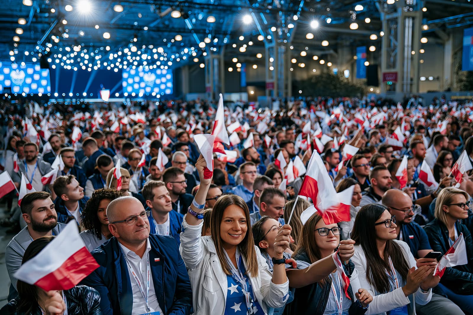 Pārpildīta izstāžu zāles publika vicina mazus karogus spilgtā pasākuma apgaismojumā AfD Magdeburgas kampaņas atklāšanas pasākumā, tālumā redzama skatuve.