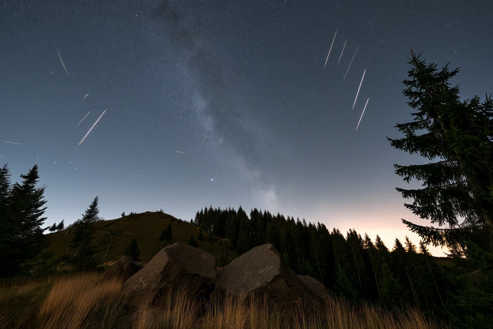 Nakts debesis virs tumša Vācijas lauku apvidus ar meteorītu svītrām un blāvu zvaigžņu joslu virs galvas.
