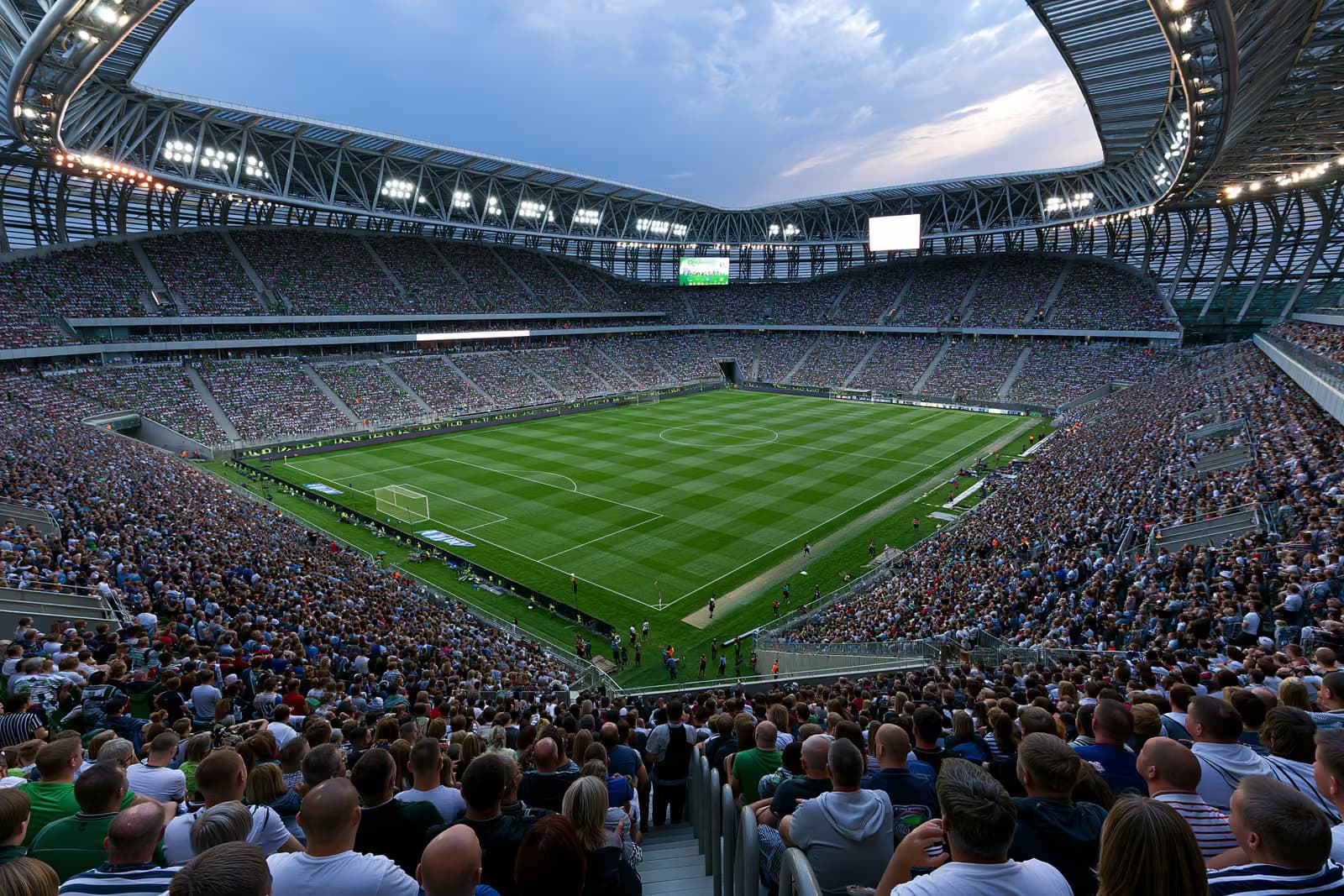 Liels futbola stadions, kas vasaras vakarā prožektoru gaismā piepildīts ar līdzjutējiem.